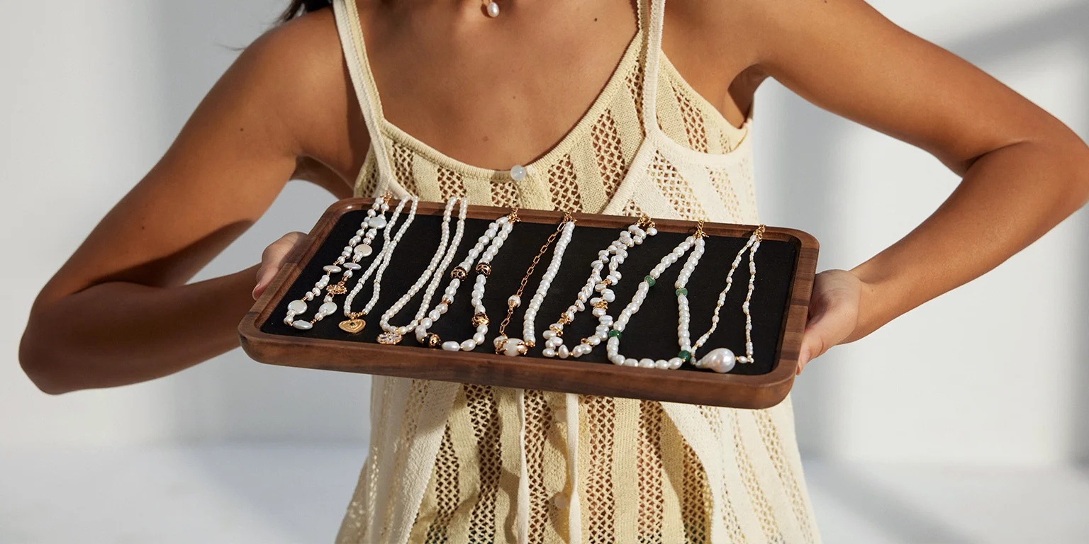 A beautiful woman is holding a square tray upon which rests an exquisite pearl necklace.
